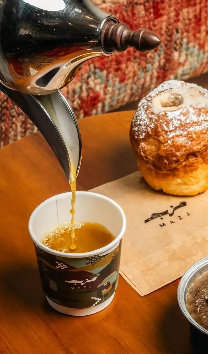 Arabic coffee poured from a dallah into a Mazj cup, next to a sugar-dusted croissant